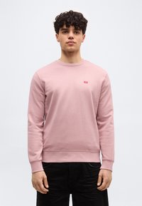 Νέος άνδρας με μπούκλες, που φοράει ροζ ανοιχτό φούτερ χωρίς κουκούλα και μαύρο παντελόνι, στέκεται μπροστά σε απλό ανοιχτό φόντο.