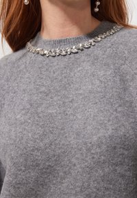 Pull en laine grise avec un col rond orné d'une rangée de pierres précieuses transparentes, ajoutant de la texture et un intérêt visuel au design.