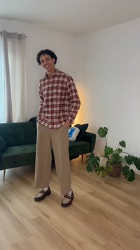 Jeune personne souriante, portant une chemise à carreaux et un pantalon beige, debout sur un sol en bois près d'un canapé vert et d'une plante en pot dans une pièce lumineuse.