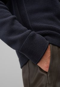 Marinblå strukturstickad tröja med ribbade ärmslut, visas tillsammans med släta bruna läderbyxor. Detalj på ärmen och handen i fickan synlig.