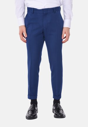 Pantaloni su misura blu realizzati in un tessuto liscio, con pieghe frontali, passanti per la cintura e una vestibilità affusolata, abbinati a mocassini neri.