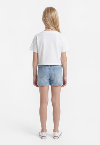 Enfant aux longs cheveux blonds portant un t-shirt blanc, un short en jean bleu clair et des baskets blanches, debout de dos sur fond blanc.