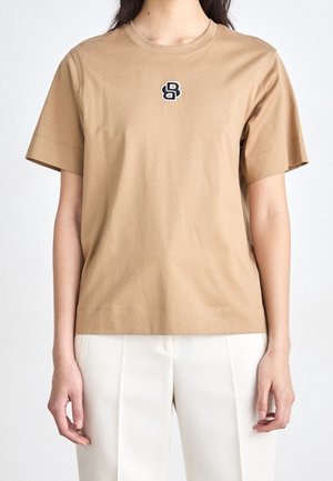 Nainen, joka pukeutunut beigeen lyhythihaiseen t-paitaan, jossa on pieni mustavalkoinen logo rinnassa, yhdistettynä valkoisiin housuihin.