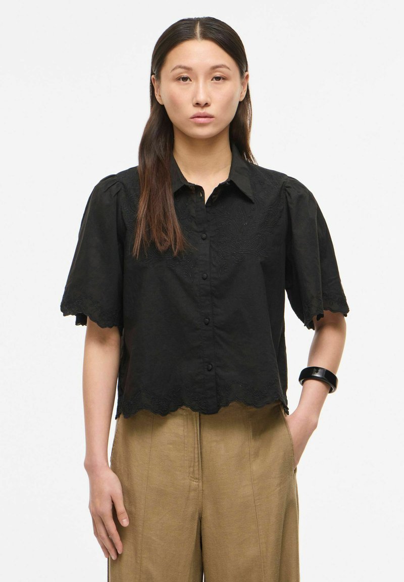 Femme aux cheveux bruns raides porte un chemisier noir à manches courtes boutonné et un pantalon beige, debout, une main dans la poche, expression neutre.