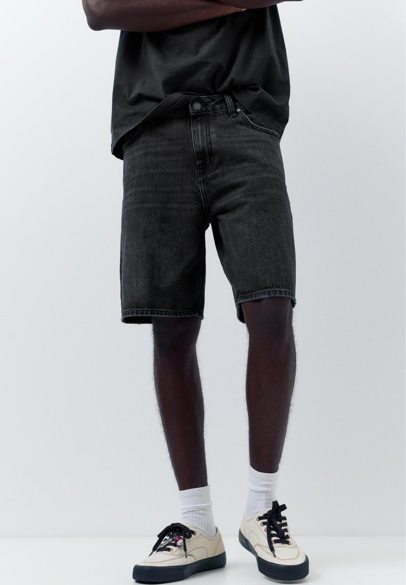Homme portant un short en jean noir, des chaussettes blanches, des baskets beiges et un t-shirt noir, debout les bras croisés devant un fond uni.