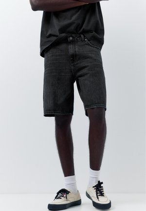 Uomo con pantaloncini in denim nero, calzini bianchi, sneakers beige e maglietta nera in piedi con le braccia incrociate su sfondo semplice.