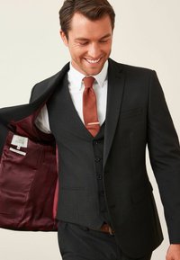 Musta puku slim fit -mallilla, jossa on viininpunainen vuori ja yhteensopiva solmio. Valkoinen kauluspaita, jota täydentää vaaleanruskea vyö.