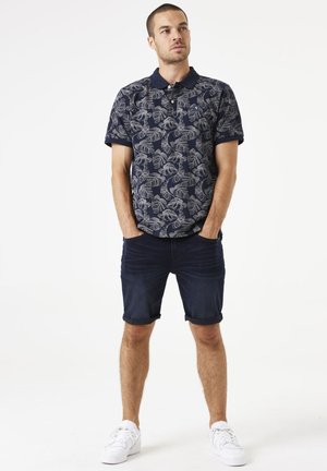 Camiseta polo navy con patrón de hojas grises, de manga corta y con cuello; combinada con pantalones cortos de denim enrollados y zapatillas blancas.