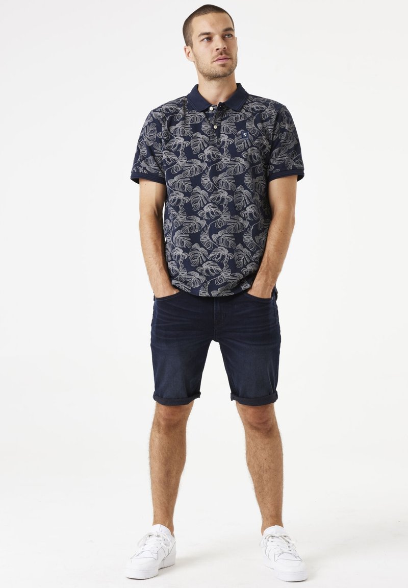 Marineblauwe poloshirt met een grijs bladpatroon, korte mouwen en kraag; gecombineerd met opgerolde denimshorts en witte sneakers.