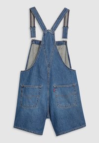 Salopette courte en jean avec bretelles ajustables, deux poches à l'arrière et détails de couture sur un fond uni clair.