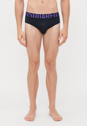 Mann trägt schwarze Boxershorts mit einem breiten lila Bündchen, das mit fetter Schrift bedruckt ist, und steht barfuß vor einem schlichten weißen Hintergrund.