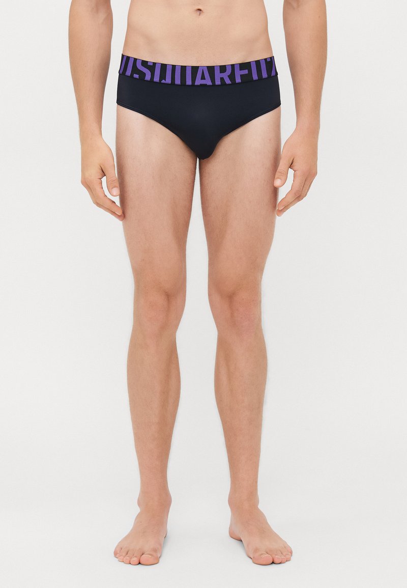 Mann trägt schwarze Boxershorts mit einem breiten lila Bündchen, das mit fetter Schrift bedruckt ist, und steht barfuß vor einem schlichten weißen Hintergrund.