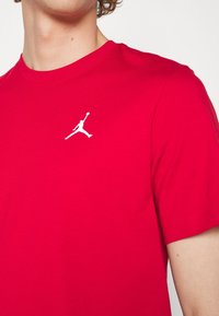 Persona llevando una camiseta roja brillante con un pequeño logo de Jumpman blanco en el lado izquierdo del pecho, visible desde los hombros hasta la parte superior del torso.