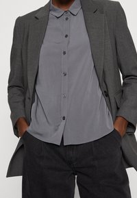 Blazer gris con solapas de muesca, combinado con una camisa de botones gris claro que presenta una textura suave y cuello corto. Pantalones oscuros.