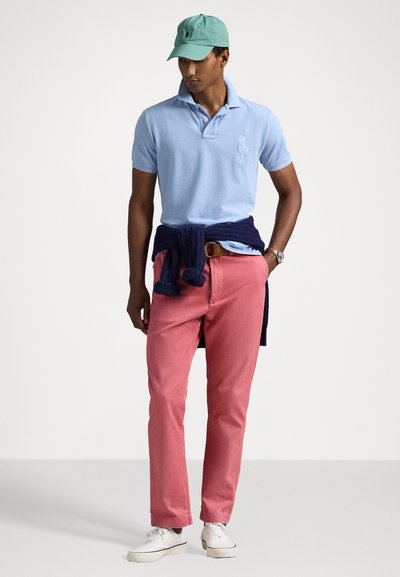 Light blue polo shirt, pink chinos, navy sweater tied at the waist, green cap, white sneakers, featuring classic fit and embroidered logo.