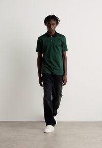 Zelené polo tričko s čiernym golierom, skombinované s tmavými džínsy a bielymi teniskami. Model stojí pred neutrálnym pozadím.