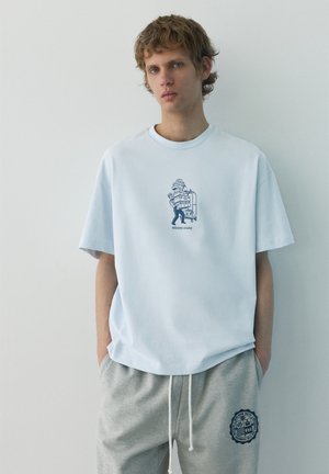 Tânăr stă lângă un perete alb, purtând un tricou bleu cu grafică de bagaje și pantaloni de trening gri cu un emblema circulară.