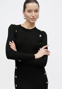 Kobieta o brązowych włosach związanych w niski kok, ubrana w czarną prążkowaną sukienkę z złotymi guzikami i złote kolczyki koła, z założonymi ramionami na białym tle.