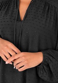 Blusa nera con pois testurizzati, scollatura a V profonda, spalle arricciate e maniche a palloncino. Tessuto liscio, maniche lunghe e dettagli sottili.