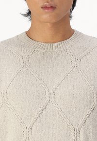 Light beige knitted sweater featuring a diamond pattern, ribbed neckline, and textured fabric. Design emphasizes clean lines and minimalism.