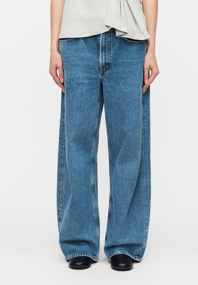 Άτομο που φοράει φαρδιά μπλε τζιν, μαύρα παπούτσια και ανοιχτό γκρι μπλουζάκι, στέκεται μπροστά σε ένα απλό λευκό φόντο.