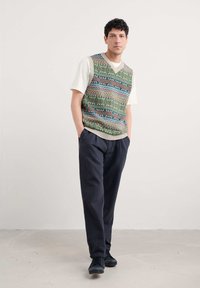 Mann steht mit den Händen in den Taschen, trägt ein weißes T-Shirt, eine bunt gemusterte Strickweste, dunkle Hosen und schwarze Sneaker vor einem einfarbigen Hintergrund.