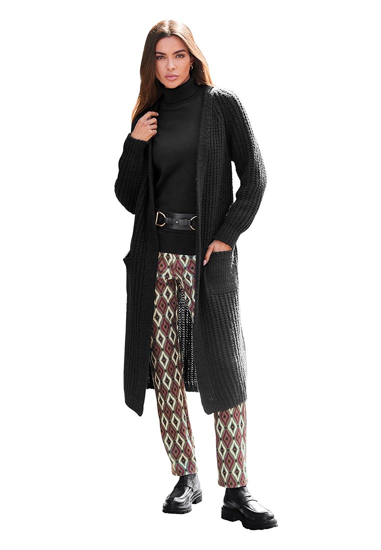 Czarny sweter z dzianiny z otwartym przodem i kieszeniami, noszony na czarnym golfie i wzorzystych spodniach w odcieniach czerwieni, zieleni i beżu.