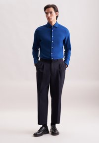 Camicia blu con bottoni, maniche lunghe, abbinata a pantaloni scuri a pieghe e scarpe eleganti nere. Tessuto liscio e vestibilità su misura.