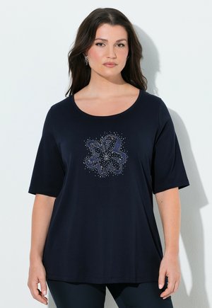 Kvinde med langt mørkt hår iført en marineblå T-shirt med korte ærmer og et centralt abstrakt blomsterprint lavet af små prikker og linjer.