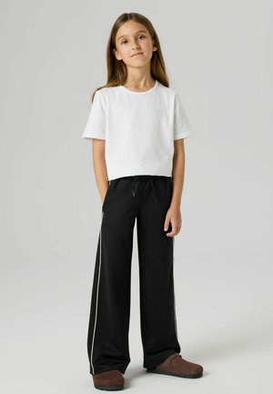 Ung person som står, iført en hvit T-skjorte, svarte vide bukser med hvite sidelister, og brune slip-on-sko, med hendene delvis i lommene.