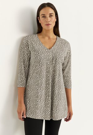 Vrouw met lang donker haar, goudkleurige oorbellen en een grijze top met V-hals en witte onregelmatige stippen, staand tegen een witte muur.