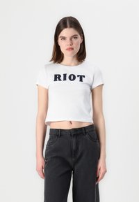 Bela kratka majica s kratkimi rokavi in napisom "RIOT" v črni barvi, nekoliko raztegljiva. Ujemajoč se s črnimi kavbojkami visoke pasu, teksturirano blago.