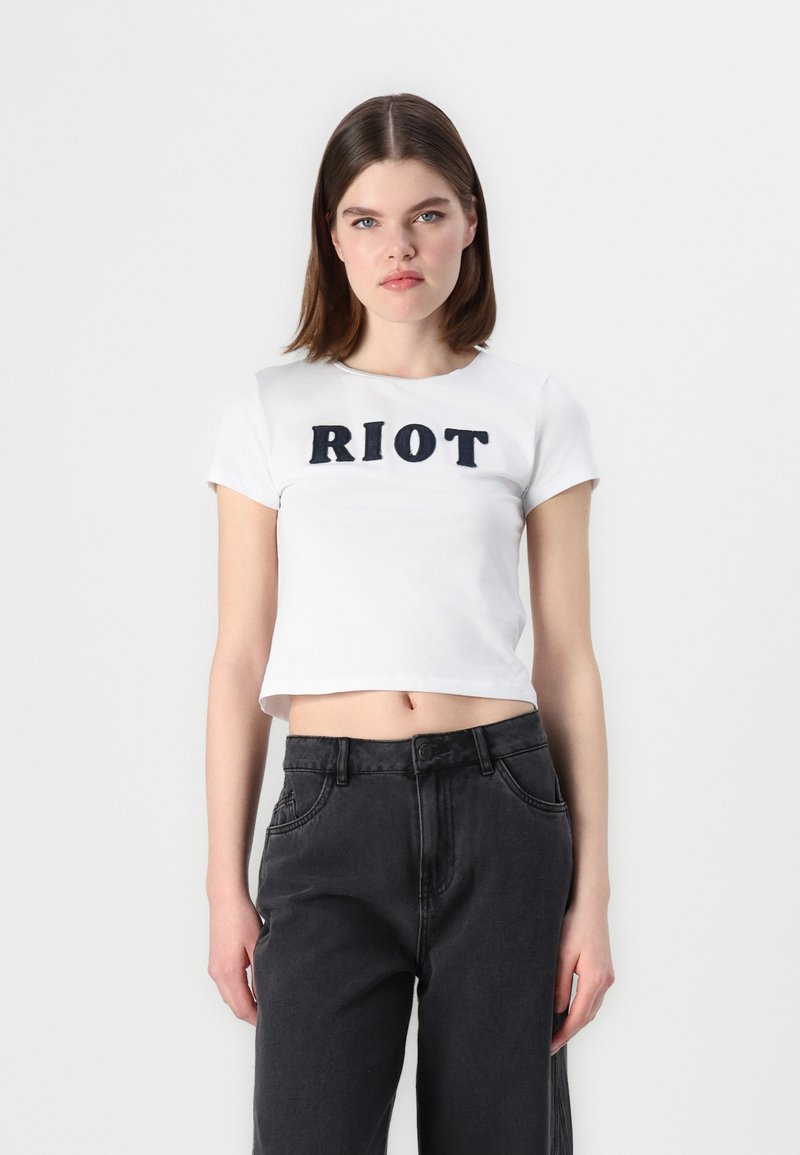 Bela kratka majica s kratkimi rokavi in napisom "RIOT" v črni barvi, nekoliko raztegljiva. Ujemajoč se s črnimi kavbojkami visoke pasu, teksturirano blago.