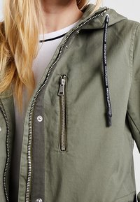Olive zelená bunda se zapínáním na zip, nastavitelnou kapucí a kontrastní vnitřní podšívkou, vybavená stříbrným kováním a stahovací šňůrkou.