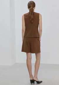 Mujer con cabello largo, vestida con una blusa marrón sin mangas de rayas finas y shorts a juego, de pie de espaldas, usando mules negros de tacón en una habitación minimalista.