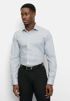 Mann trägt ein hellgraues, geknöpftes Hemd, das in schwarze Hosen mit schwarzem Gürtel gesteckt ist, steht vor einem weißen Hintergrund und schaut nach links.