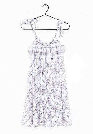 Robe sans manches à carreaux blancs et gris avec des bretelles ajustables nouées, suspendue sur un cintre noir sur fond blanc.