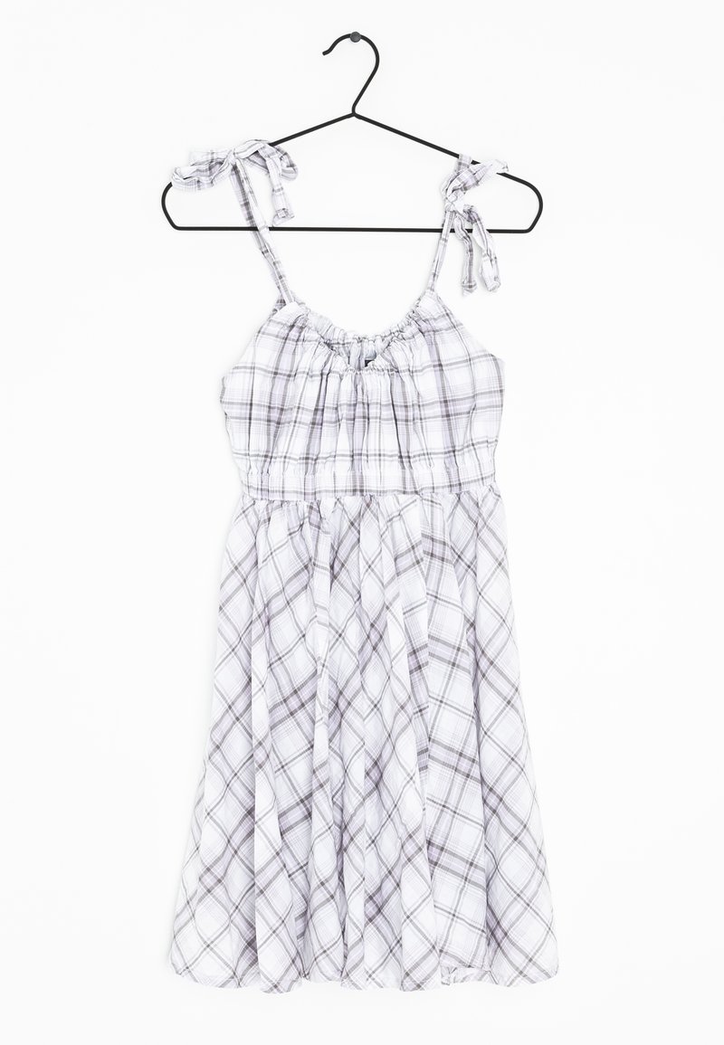 Robe sans manches à carreaux blancs et gris avec des bretelles ajustables nouées, suspendue sur un cintre noir sur fond blanc.
