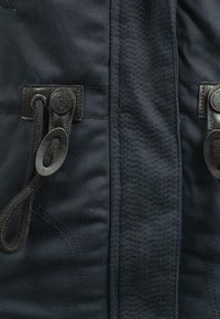 Mörkblå jacka med en slät finish, försedd med två svarta dragsnören, synliga sömmar och en quiltad textur längs kanterna.