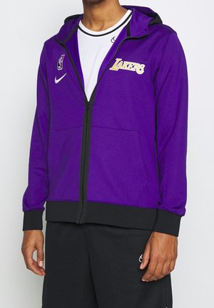 Mężczyzna w fioletowej bluzie z kapturem Lakers na zamek, z czarnymi mankietami i ściągaczem, pod spodem biała koszulka Nike i czarne spodnie Nike.