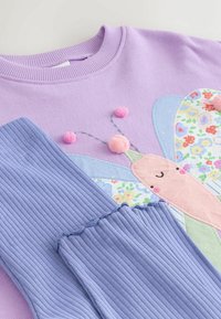 Hanorac mov cu design cu fluture, având pete roz, albastre și florale, asociat cu manșete din bumbac albastru deschis și accente roz cu pom-pom.