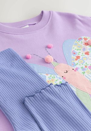 Fioletowa bluza z motywem motyla, ozdobiona różowymi, niebieskimi i kwiatowymi naszywkami, wyposażona w prążkowane, jasnoniebieskie mankiety oraz różowe pomponiki.