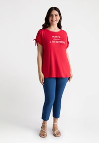Top corto rojo de manga corta con mangas atadas y texto blanco, combinado con pantalones cortos azules y sandalias beige. Tela suave, corte relajado.