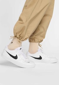 Białe sneakersy Nike z czarnym logo, w połączeniu z jasnobeżowymi zwężanymi spodniami i beżowymi, teksturowanymi skarpetkami, stojące na neutralnym tle.