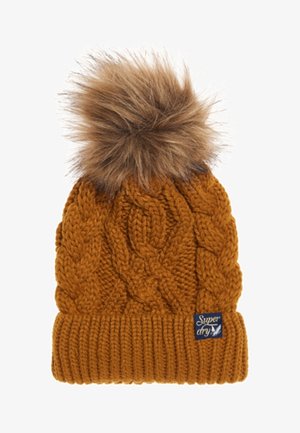 Bonnet tricoté jaune moutarde avec motifs torsadés, bord-côte, pompon en fausse fourrure marron et petite étiquette de marque rectangulaire sur le bord.