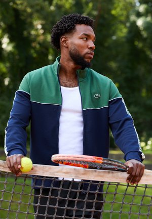 Mann i grønn og marineblå jakke som holder tennisball og racketer, lent mot tennisnettet utendørs med trær i bakgrunnen.