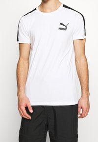 Vit kortärmad T-shirt i mjukt tyg. Svarta axelstrimmor och logotypdetalj på bröstet. Rund halsringning och avslappnad passform.