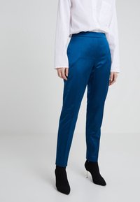 Bright blue tailored pants with a smooth texture and fitted shape, paired with black sock-style heels. White button-up shirt visible above.
