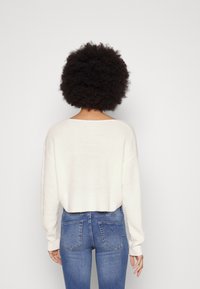 Pull blanc en tricot avec une coupe ample, un col large et des manches longues, associé à un jean bleu ajusté. La texture semble douce et confortable.