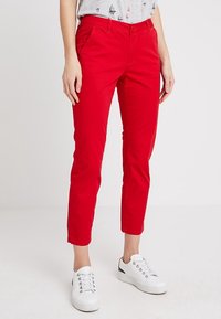 Pantalon rouge en coton coupe slim avec poches latérales et coutures contrastantes. Associé à des baskets blanches, présenté sur un fond neutre.